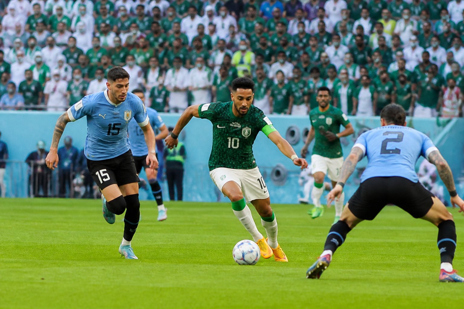 Suudi Arabistan ve Uruguay takımları arasında Dünya Kupası maçında futbolcular sahada mücadele ediyor, tribünler dolu ve coşkulu.