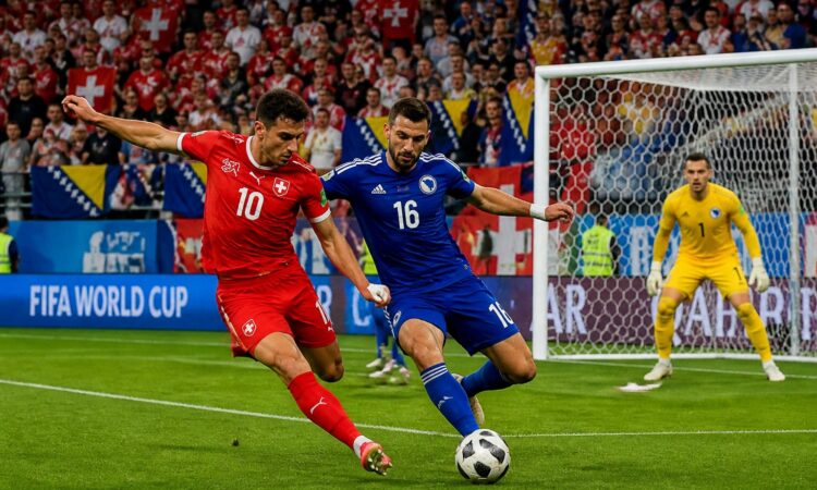 İsviçre ve Bosna Hersek takımları arasında Dünya Kupası maçında iki futbolcu top için mücadele ediyor, kalenin önünde yoğun bir atmosfer var.