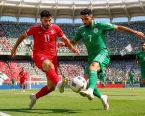 Ürdün ve Cezayir milli takımları arasında bir futbol maçında, iki oyuncu top için mücadele ediyor, büyük bir stadyumda taraftarlar tribünde yer alıyor.