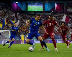 Bosna Hersek ve Katar takımlarının maç yaptığı, dolu bir stadyumda futbolcuların mücadele ettiği bir sahne.