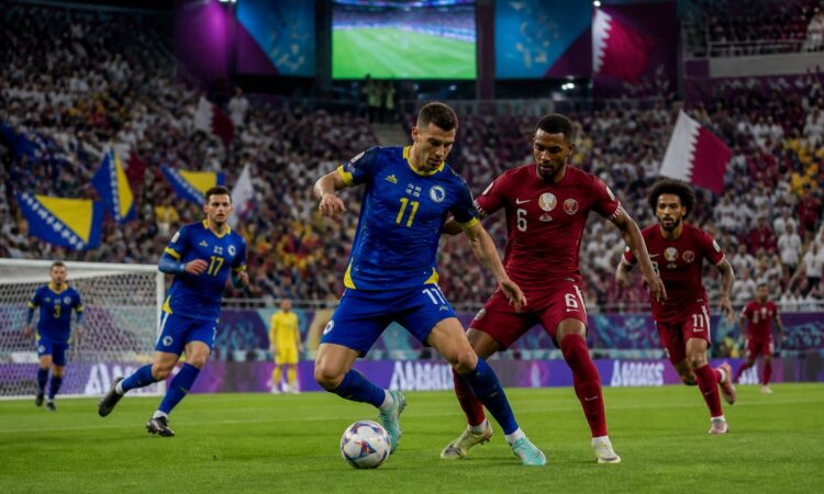 Bosna Hersek ve Katar takımlarının maç yaptığı, dolu bir stadyumda futbolcuların mücadele ettiği bir sahne.