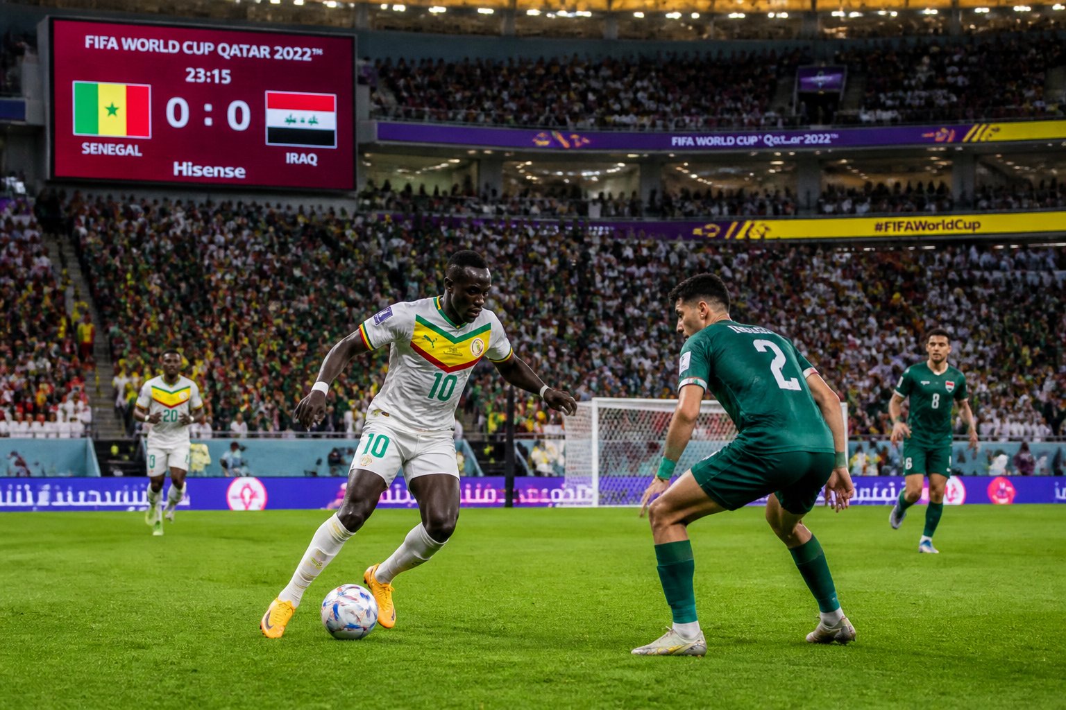 Senegal ve Irak takımları arasında oynanan bir Dünya Kupası maçında, dolu stadyumda futbolcular top için mücadele ediyor.