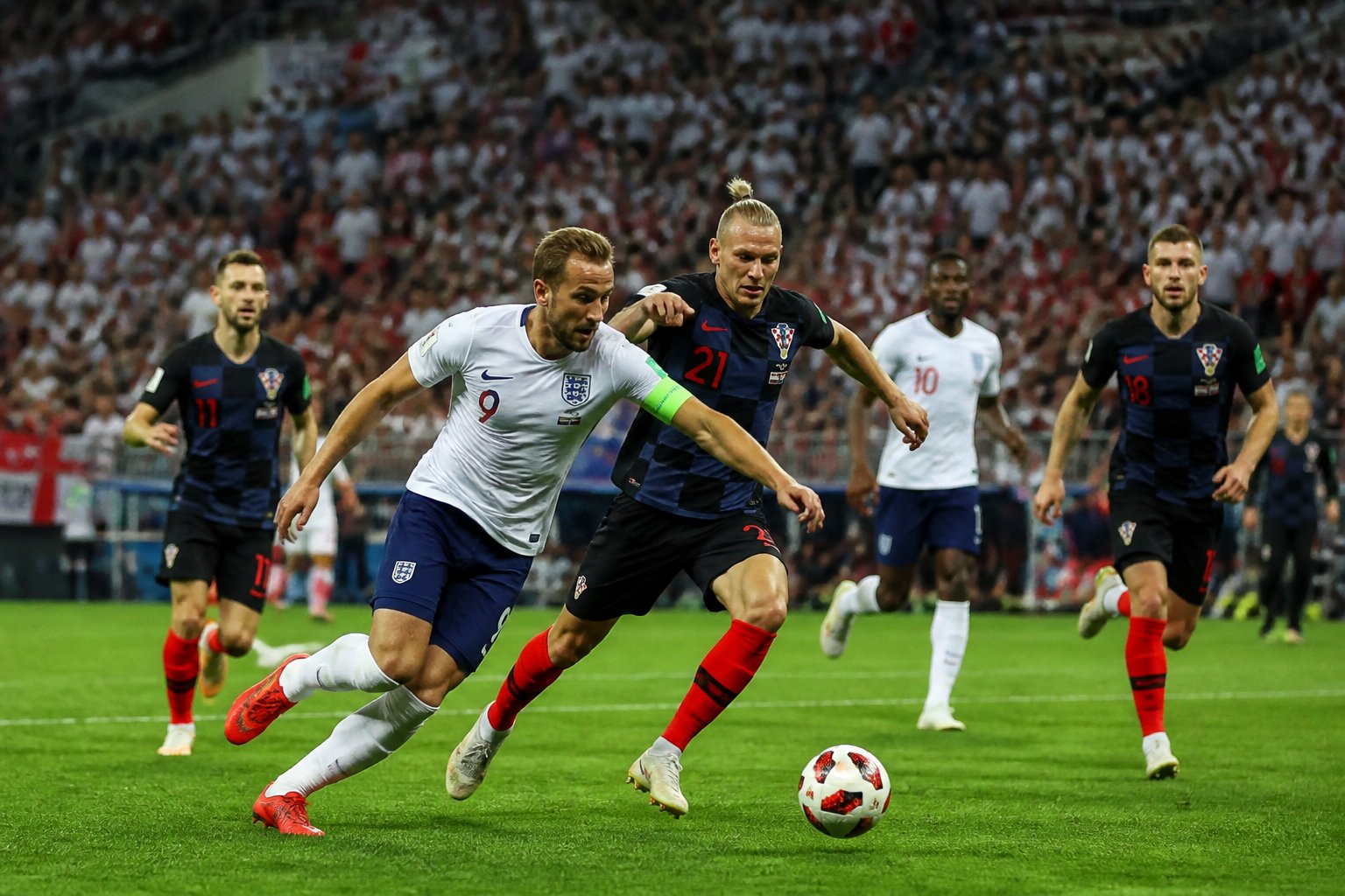 İngiltere ve Hırvatistan takımlarının Dünya Kupası maçında sahada mücadele eden futbolcuları ve dolu tribünleri gösteren bir futbol maçı sahnesi.