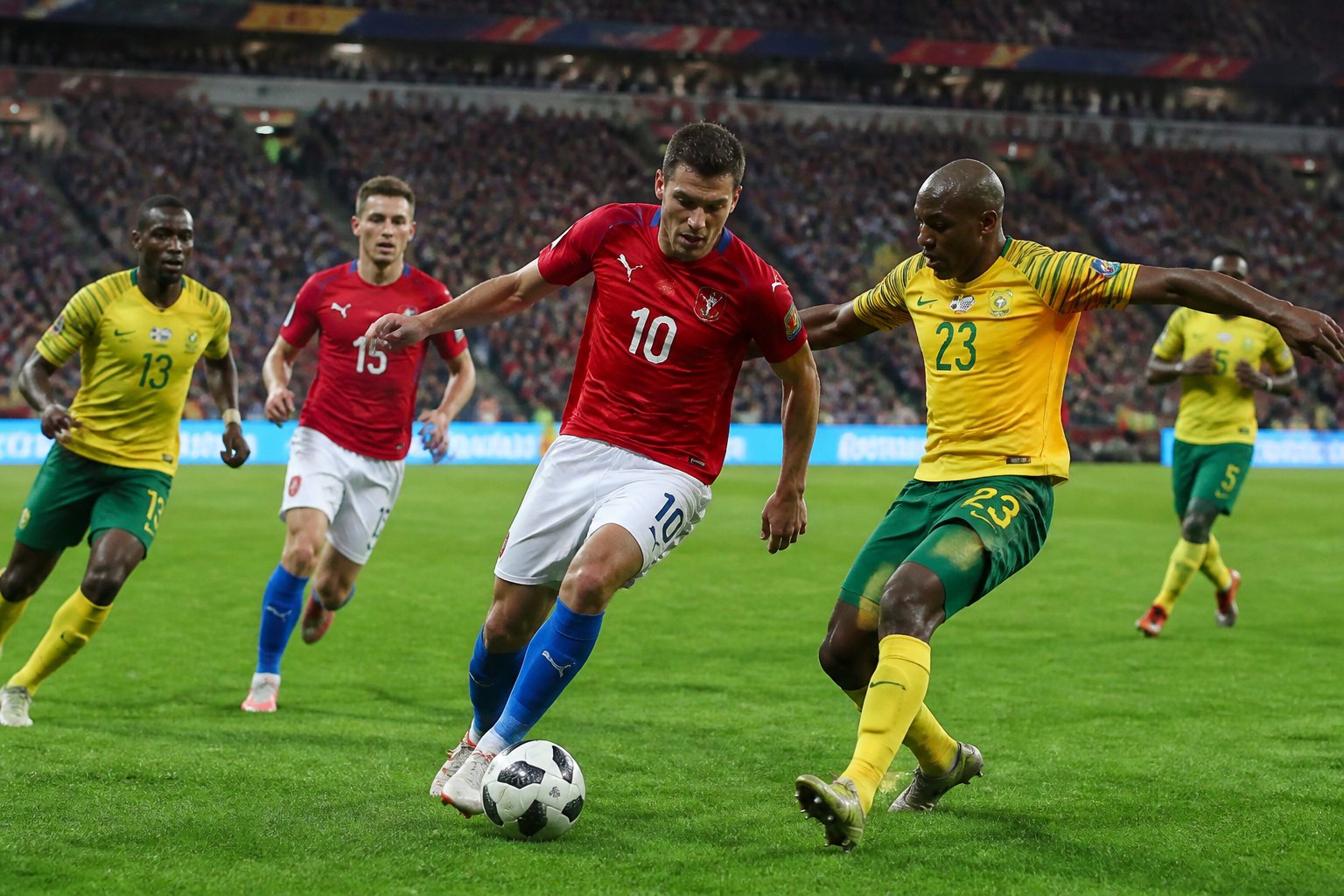 Çek Cumhuriyeti ile Güney Afrika arasında bir Dünya Kupası futbol maçı sırasında sahada mücadele eden oyuncular ve tribünlerde tezahürat yapan taraftarlar.