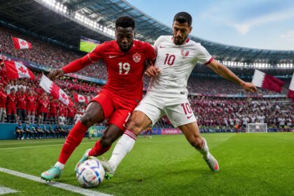 Kanada ve Katar takımları arasında bir Dünya Kupası maçında, sahada top için mücadele eden iki futbolcu ve tribünde taraftarlar.